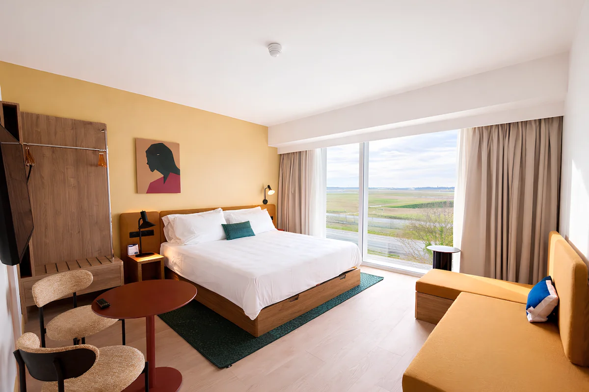 Chambre de l'hôtel Hampton by Hilton à Mérignac : lit double, mur jaune, banquette et large baie vitrée ouvrant sur la campagne
